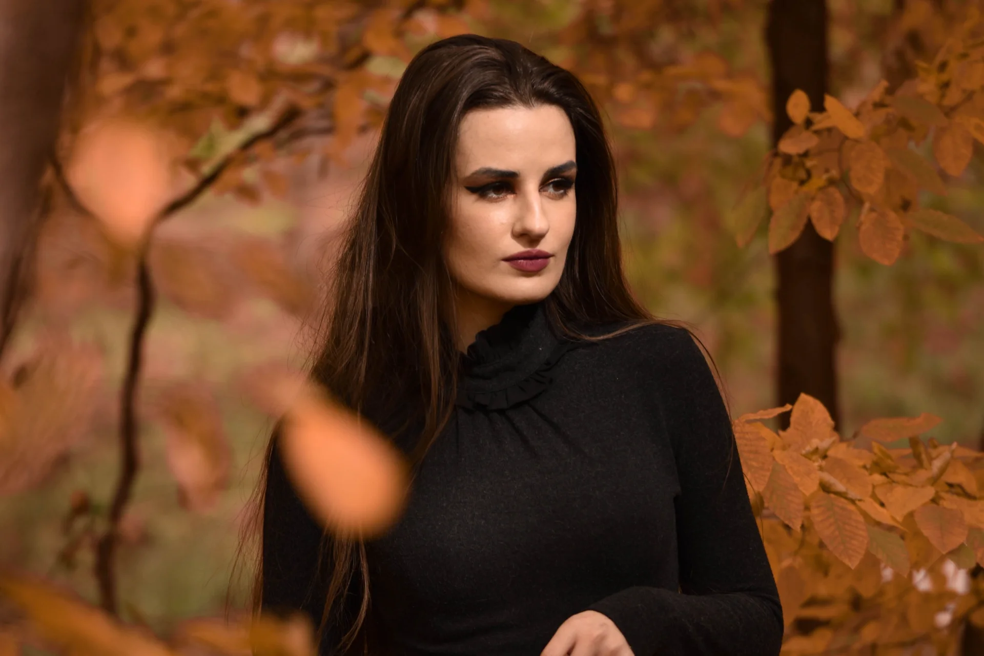 Młoda kobieta w eleganckiej czarnej sukience pozuje w jesiennym lesie, otoczona drzewami.